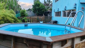 A rectangular wooden pool with a metal ladder, surrounded by lush greenery and a blue building in the background. - L'Althéa Bleu, 2 minutes from the sargasso-free beach  (Sainte-Rose)