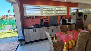 A compact kitchen with a dining area, featuring a table with a red tablecloth, wicker chairs, and a red sofa. - L'Althéa Bleu, 2 minutes from the sargasso-free beach  (Sainte-Rose)