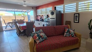 A living room with a sofa, chairs, a dining table, and a kitchen area with appliances. - L'Althéa Bleu, 2 minutes from the sargasso-free beach  (Sainte-Rose)