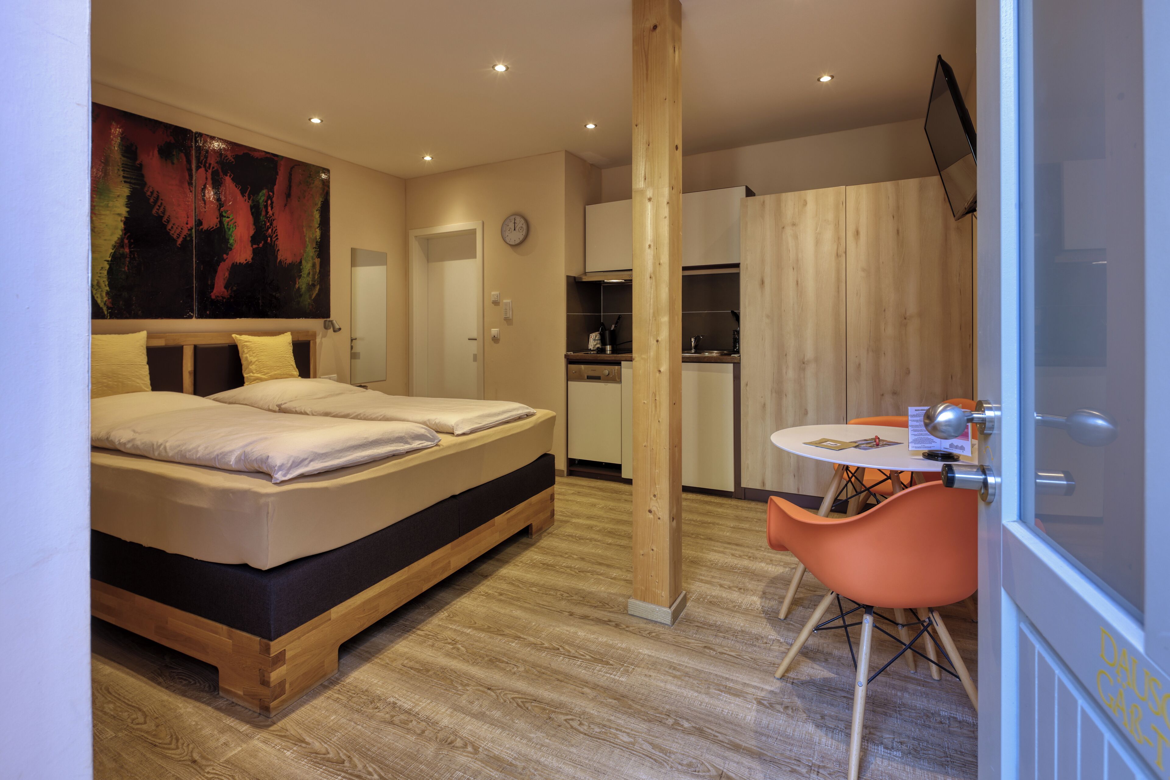 Ein modernes Schlafzimmer mit einem hölzernen Bett, einem großen abstrakten Gemälde und einer kleinen Küchenzeile mit Holzschränken und einem runden Tisch.