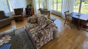 A spacious living room with a large floral sofa, a wooden coffee table, and a dining table with chairs. There are large windows providing a view of the outdoors. - Behie House - charming home on the Strait of Canso (Sand Point)