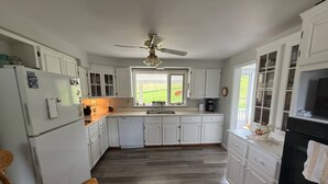 A spacious kitchen with white cabinets, a built-in oven, and a large window providing a view of the outdoors. - Behie House - charming home on the Strait of Canso (Sand Point)
