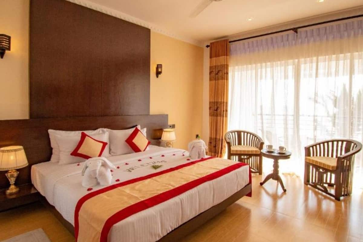 A hotel room with a large bed, two bedside tables, a small table with chairs, and a view of the outdoors through large windows.
