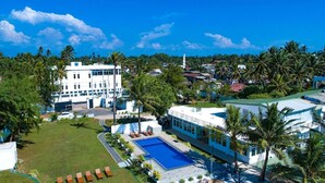 A large white building with a swimming pool and lounge chairs, surrounded by palm trees and other greenery. - Manara Beach Resort  (Beruwala)