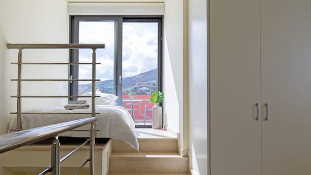 A bedroom with a bed, a window, a staircase, and a view of a red building. - Villa Misemeli (Agia Pelagia)