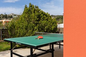 Une table de ping-pong avec un filet au milieu, entourée de verdure et avec un bâtiment en arrière-plan.