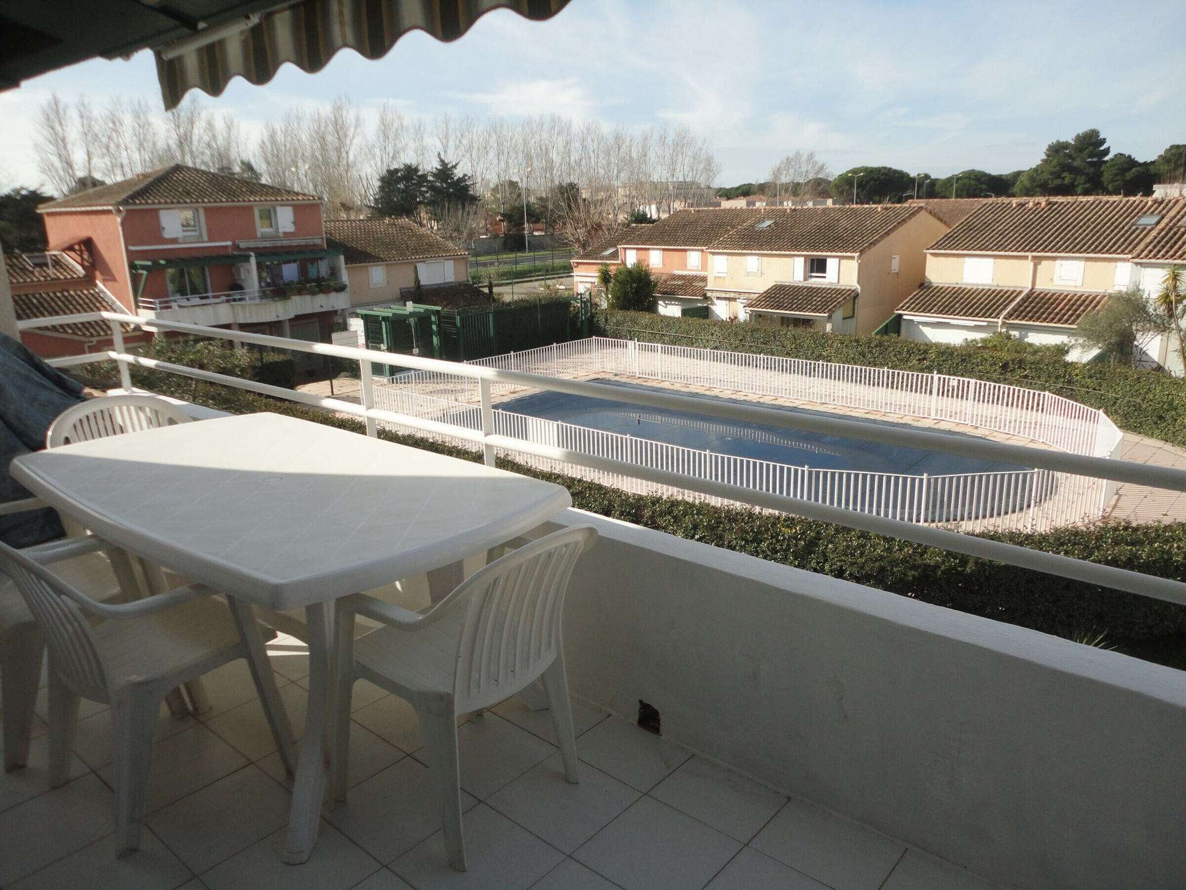 Een balkon met een witte tafel en stoelen, met uitzicht op een zwembad en woongebouwen.