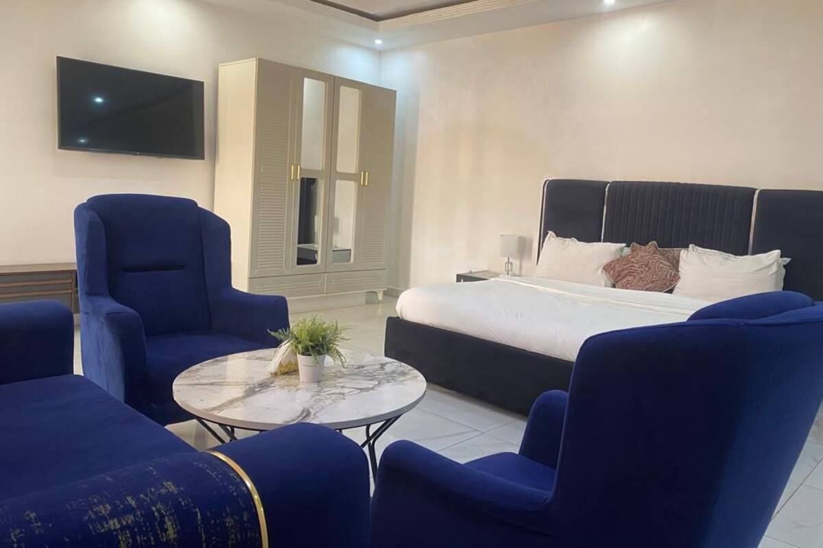 Una habitación de hotel moderna con una cama, dos sillones azules, una mesa de centro redonda con una planta, un armario y un televisor de pantalla plana.