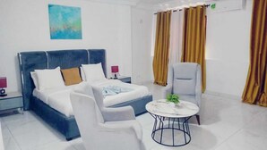 Een moderne hotelkamer met een bed, twee witte fauteuils, een kleine ronde tafel met een plant en een groot raam met gordijnen.