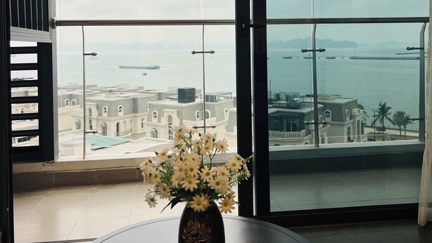 Un balcón con vista al mar y edificios, un florero con flores sobre una mesa.