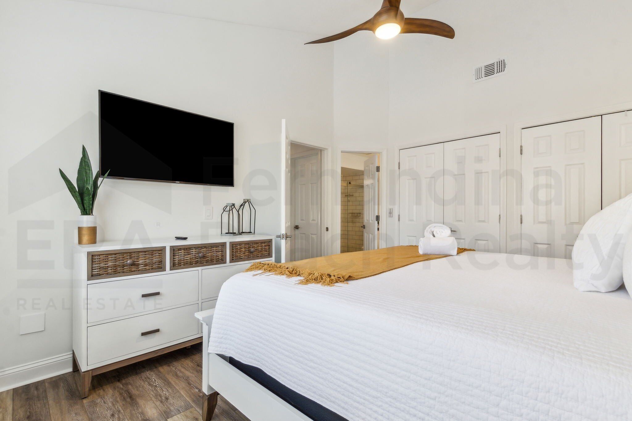 A bedroom with a bed, a TV mounted on the wall, a ceiling fan, and a wardrobe.