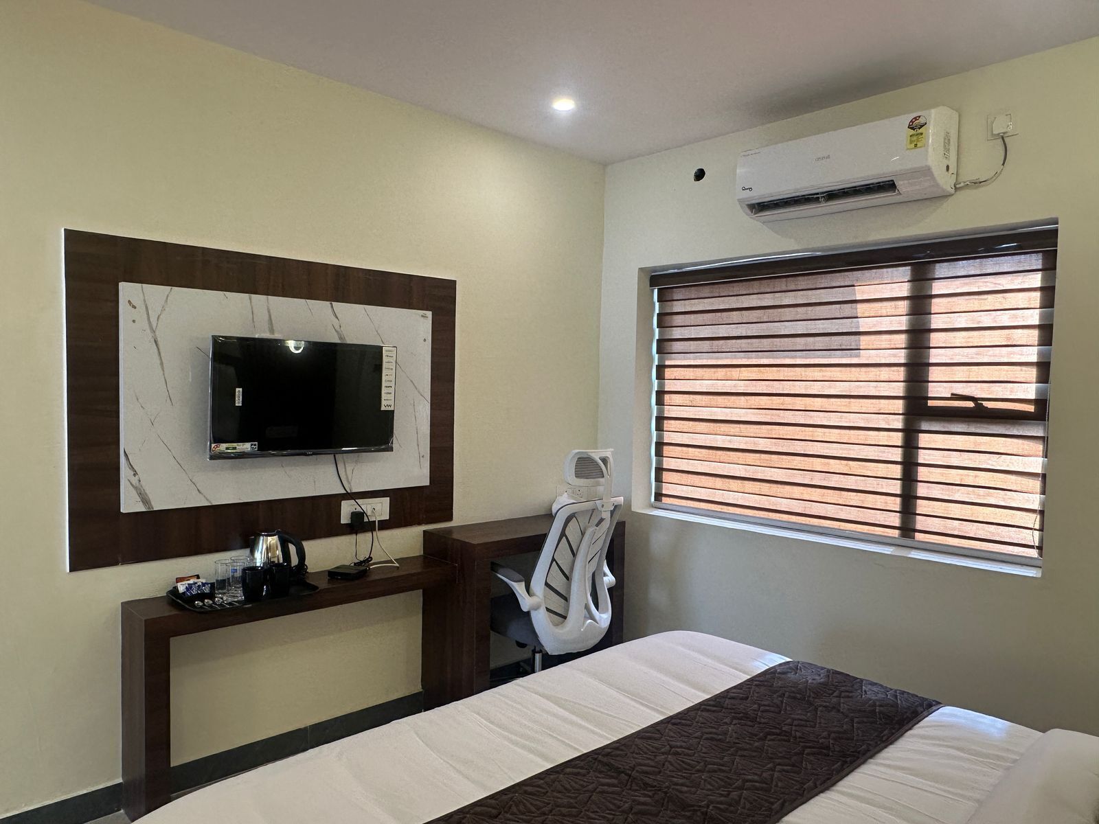 A hotel room with a bed, a TV mounted on a marble wall, a desk with a chair, and an air conditioning unit.