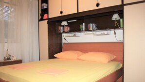 A bedroom with a bed, two pillows, a nightstand, a lamp, and a shelf with books and a small cage.