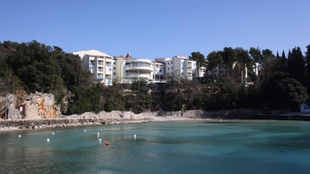 A coastal hotel with multiple buildings, a clear blue sky, and a calm sea with buoys.