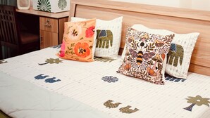 A neatly made bed with patterned pillows, a wall with decorative wall art, and a desk with a vase and a box.
