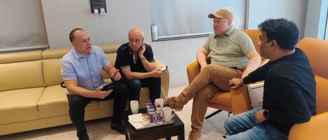 A group of four people seated on a beige couch and an orange chair in a room with a tiled floor, a small table with beverages, and a wall-mounted air conditioning unit.