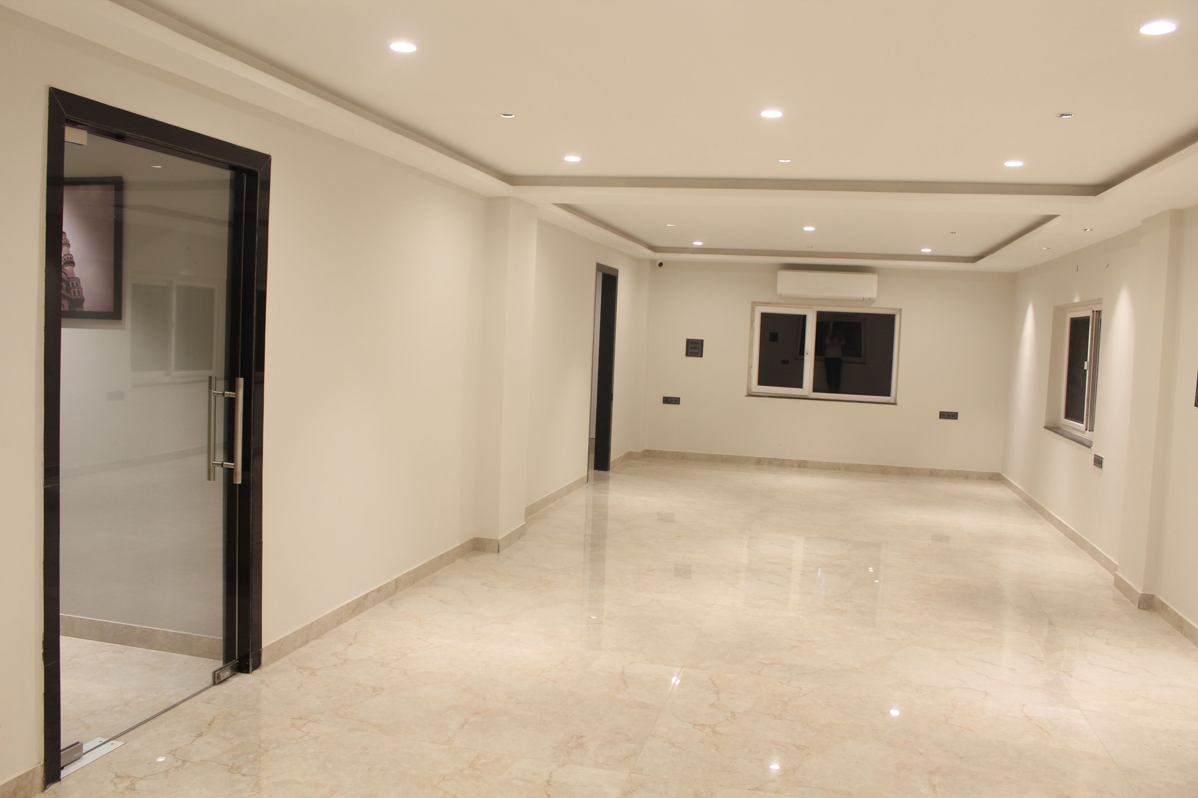 A spacious hallway with a polished marble floor, recessed ceiling lights, and a glass door on the left.
