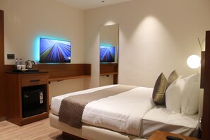 A hotel room with a bed, a flat-screen TV, a mirror, a bedside table with a remote control, and a minibar. - The Vantage Inn (Hyderabad)