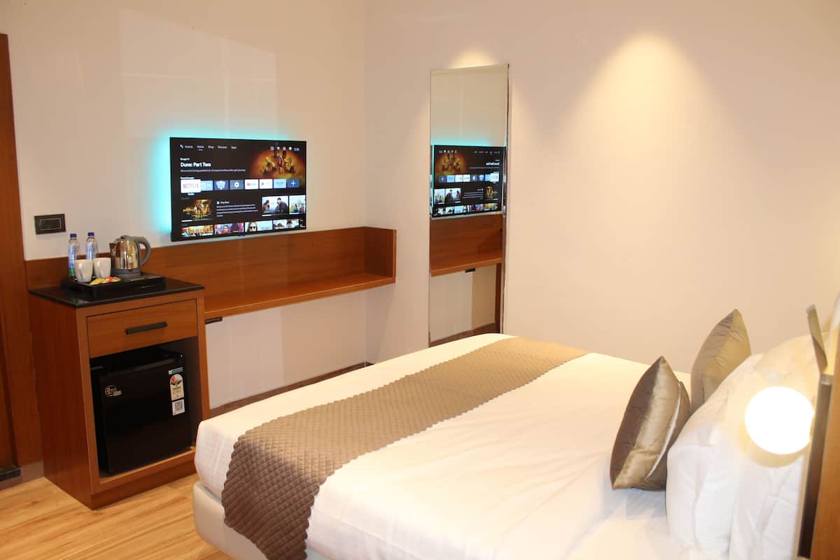 A hotel room with a bed, a wooden nightstand with a coffee maker, a flat-screen TV mounted on the wall, and a small refrigerator.