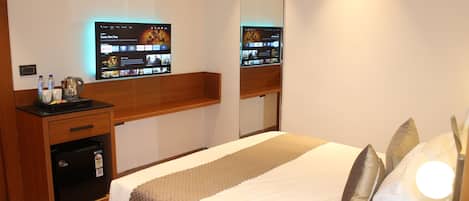 A hotel room with a bed, a wooden nightstand with a coffee maker, a flat-screen TV mounted on the wall, and a small refrigerator.