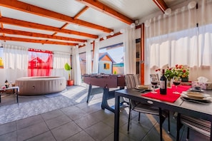 Une pièce spacieuse avec un baby-foot, un coin repas agrémenté d’un chemin de table rouge et une vue sur une maison, visible à travers de grandes fenêtres.