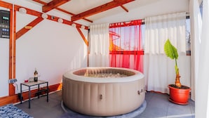 Una habitación con un jacuzzi, una planta en maceta, una mesita con una botella y vasos, y un ventanal con cortinas rojas.