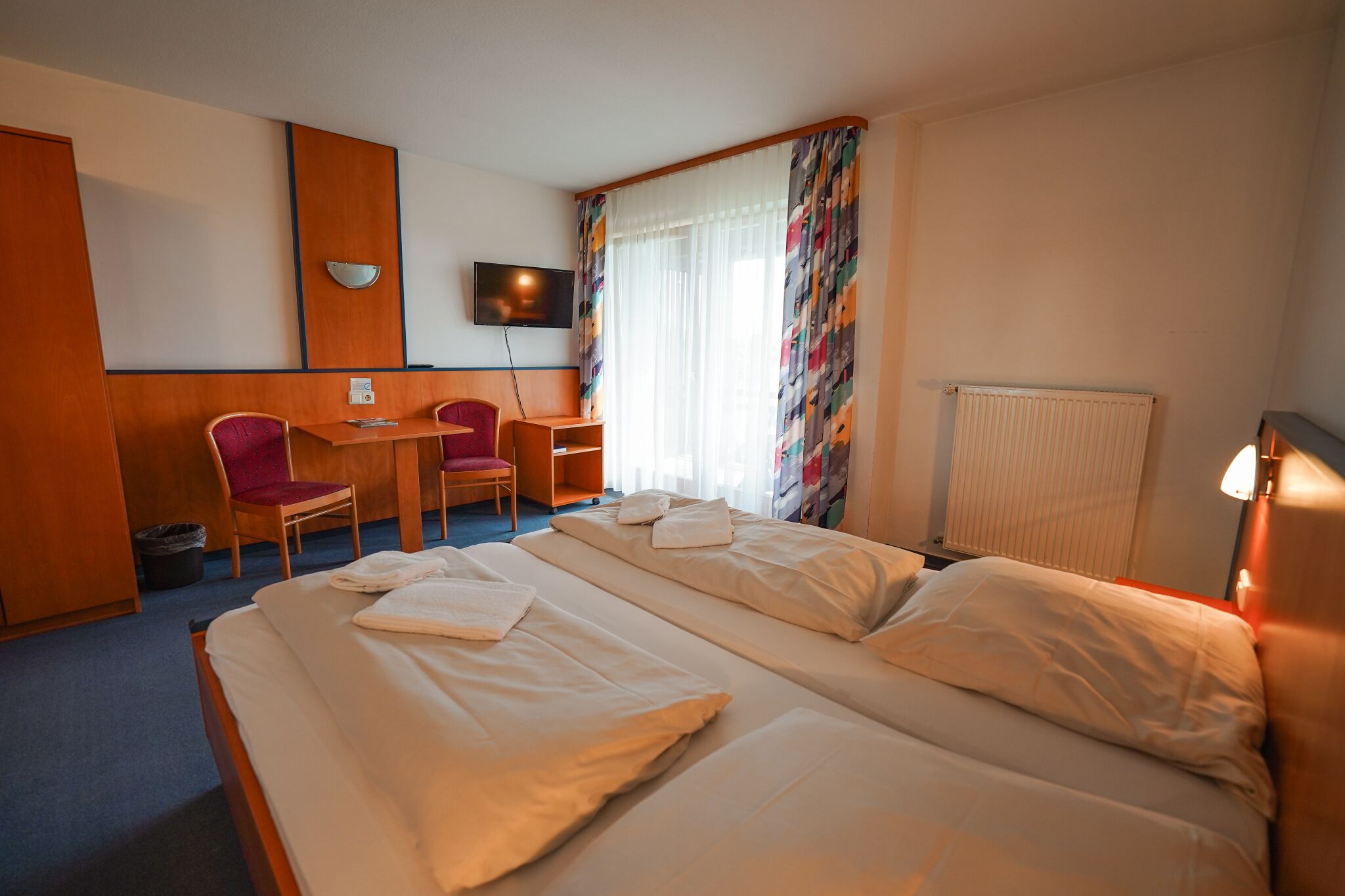 Ein Hotelzimmer mit zwei Betten, einem Schreibtisch, einem Stuhl, einem Fernseher und einem Fenster mit Vorhängen.