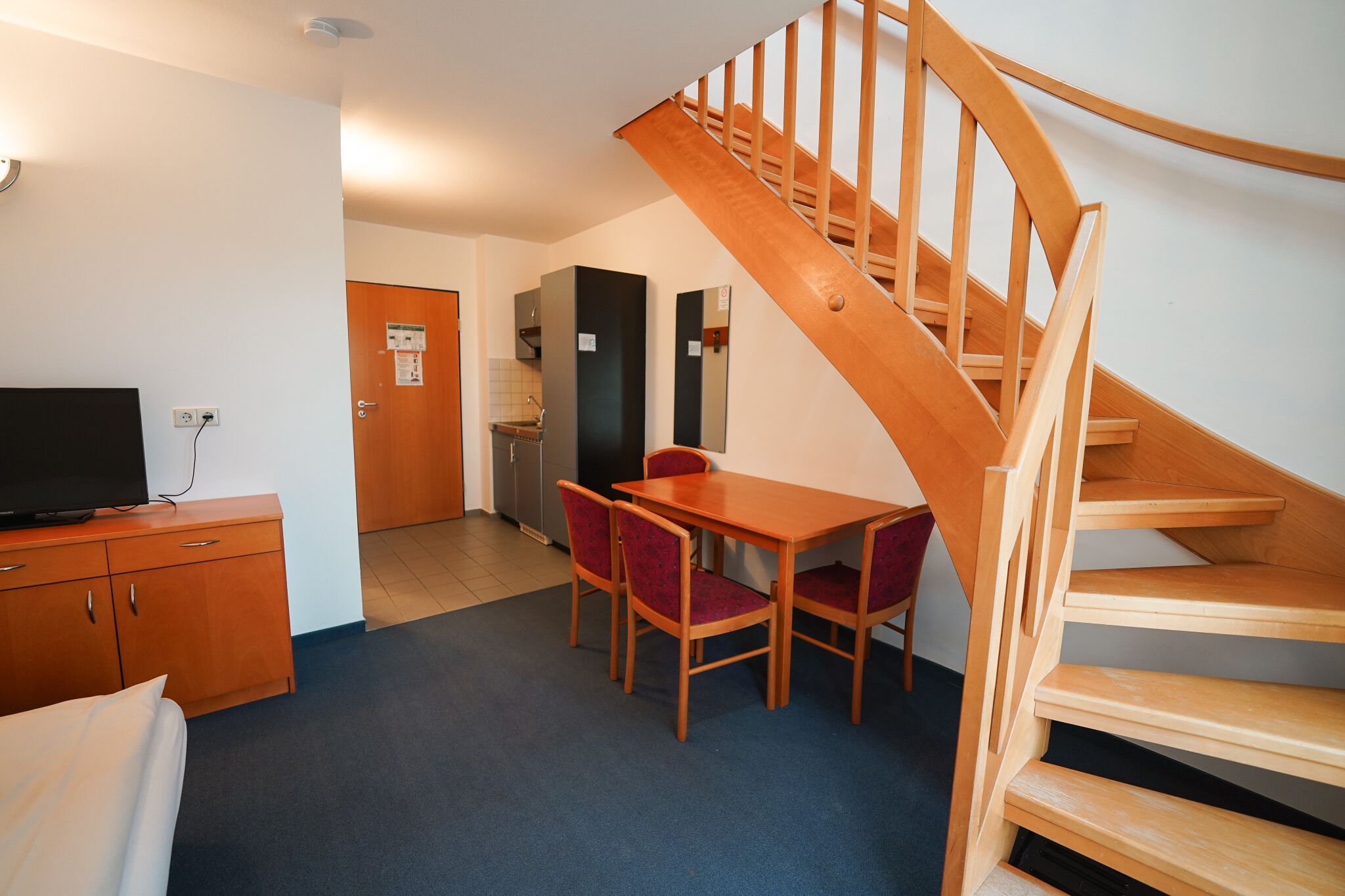 Quarto de hotel com escada de madeira, área de refeições com mesa e cadeiras, televisão em um armário de madeira e cozinha visível por uma porta aberta.