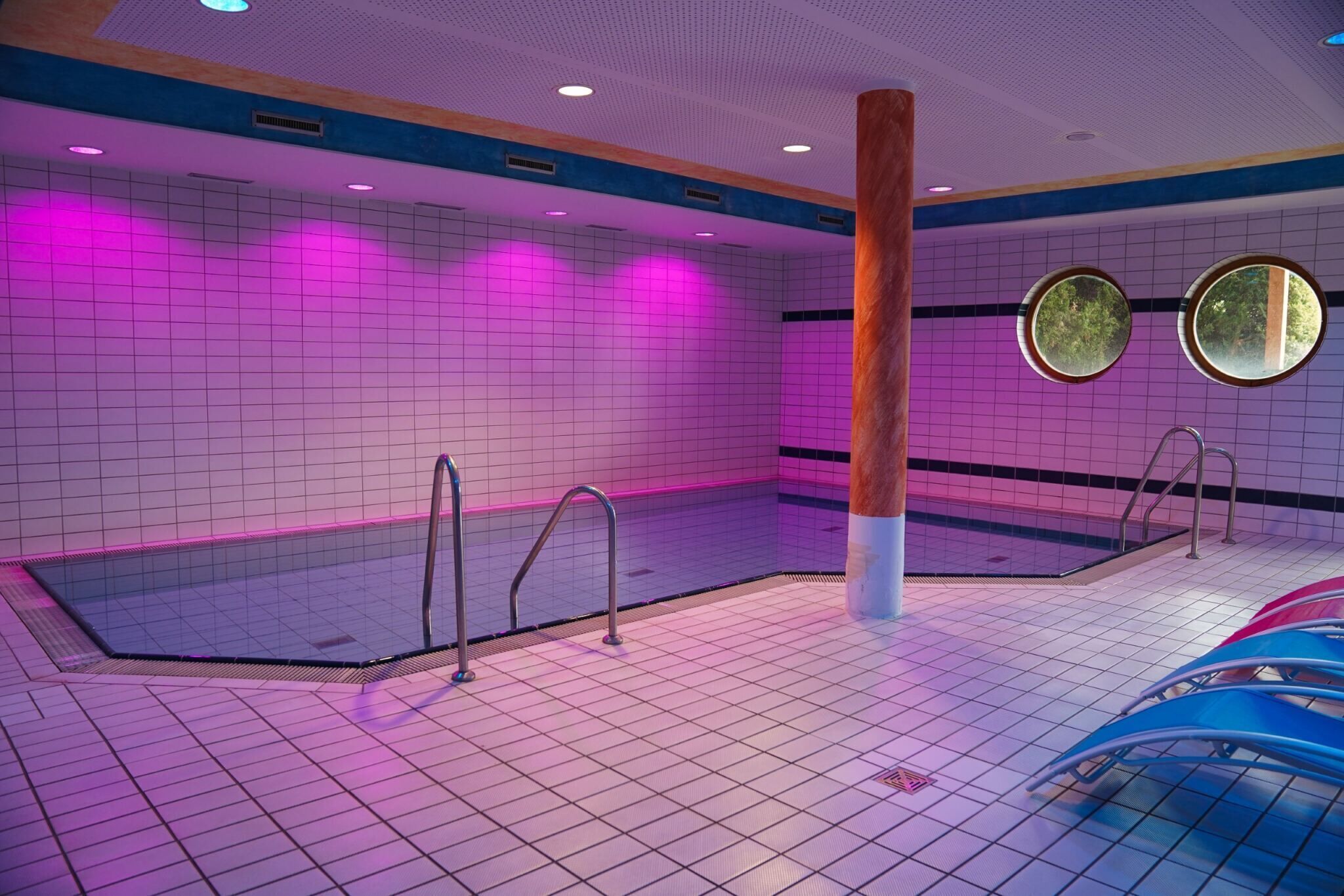 Una piscina con pareti piastrellate e decorazioni nei colori blu e bianco, dotata di due oblò rotondi e ringhiere in metallo.