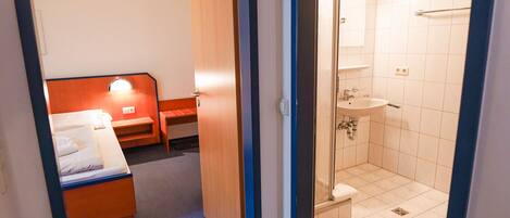 A narrow hallway leading to a bedroom with a bed and a desk, and a bathroom with a shower and sink.