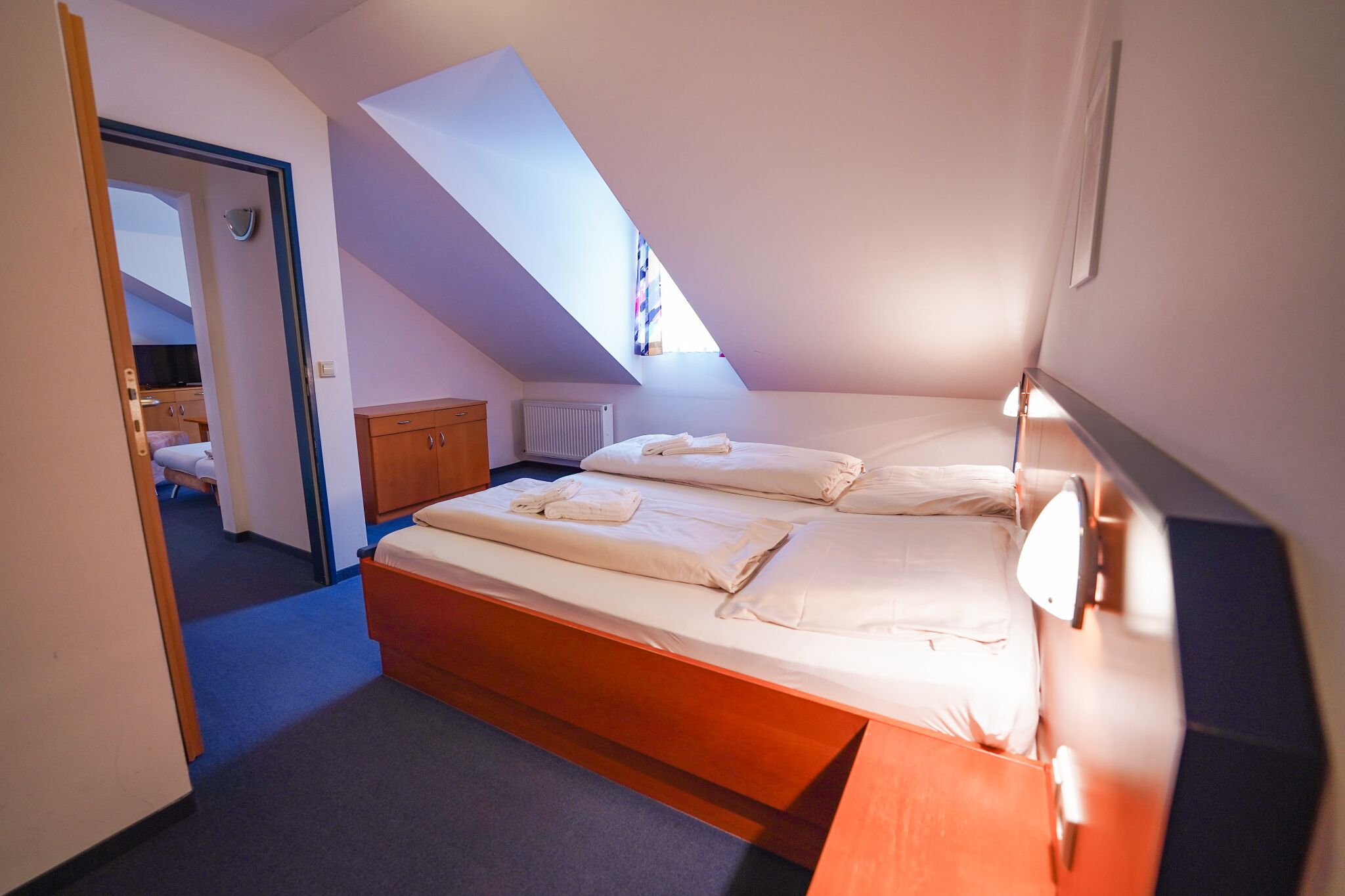 A small, well-lit bedroom with a sloped ceiling, a wooden bed frame, and a bedside table.