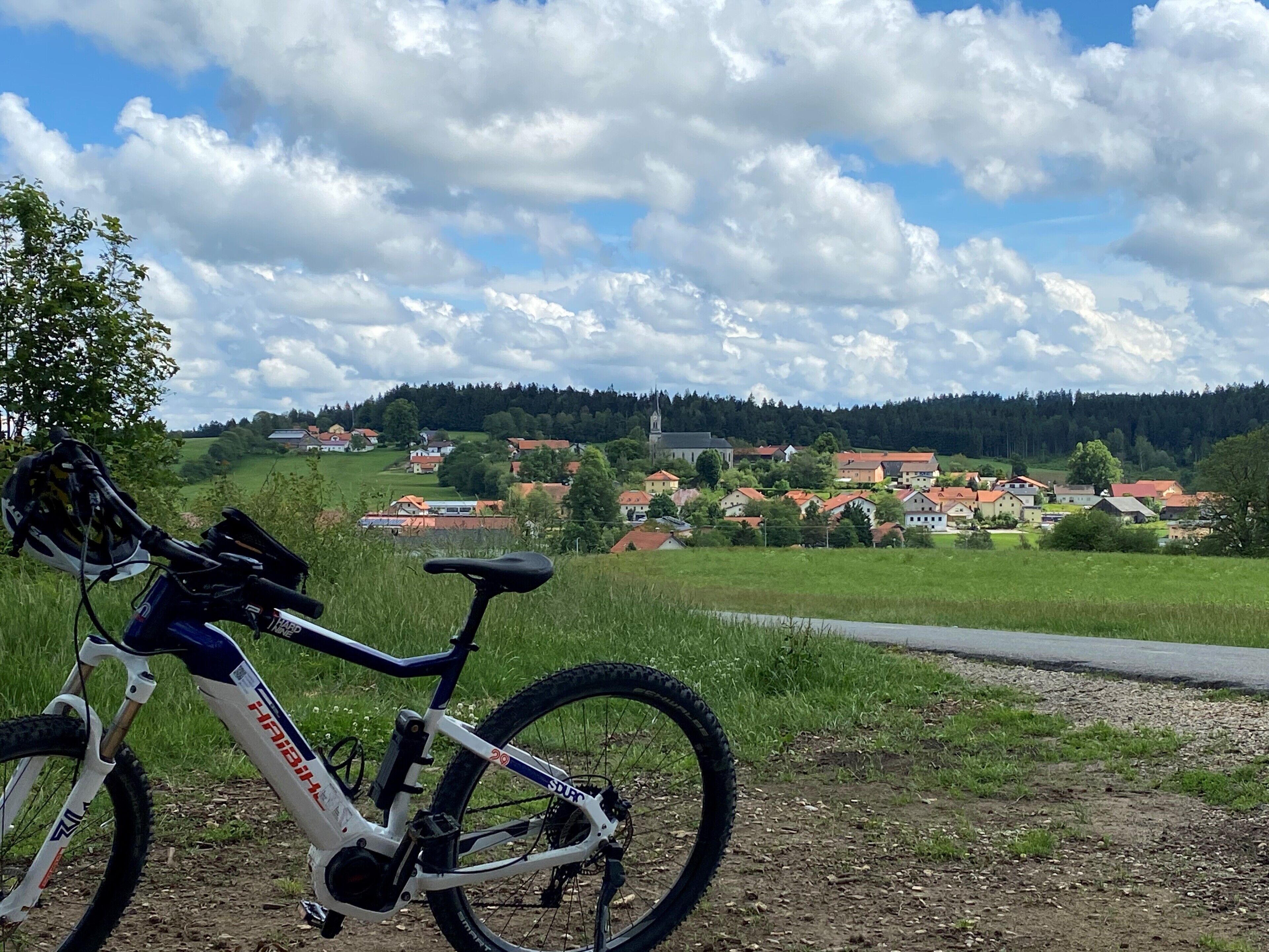 Un vélo tout-terrain est garé sur un chemin de terre, offrant une vue sur un village et des collines boisées en arrière-plan.
