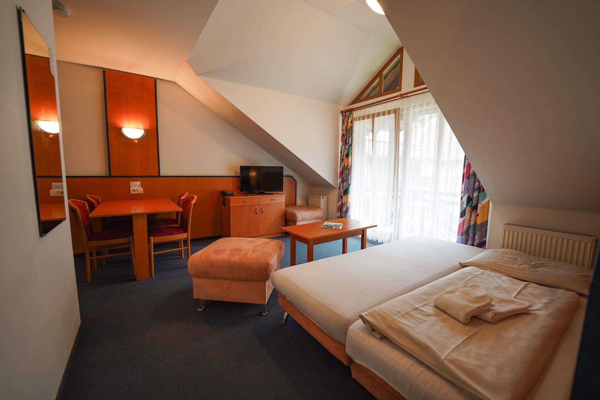 A hotel room with a bed, a desk with a chair, a television, and a window with curtains.