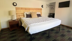 Une chambre d’hôtel avec une tête de lit en bois, un lit recouvert d’une couette blanche, une table de chevet avec une lampe, un téléviseur posé sur un meuble et un porte-manteau fixé au mur.