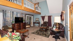 A spacious living room with a high ceiling, a wooden staircase, and a large television. - Lambs Landing Huron Family Vacation Home (Huron)