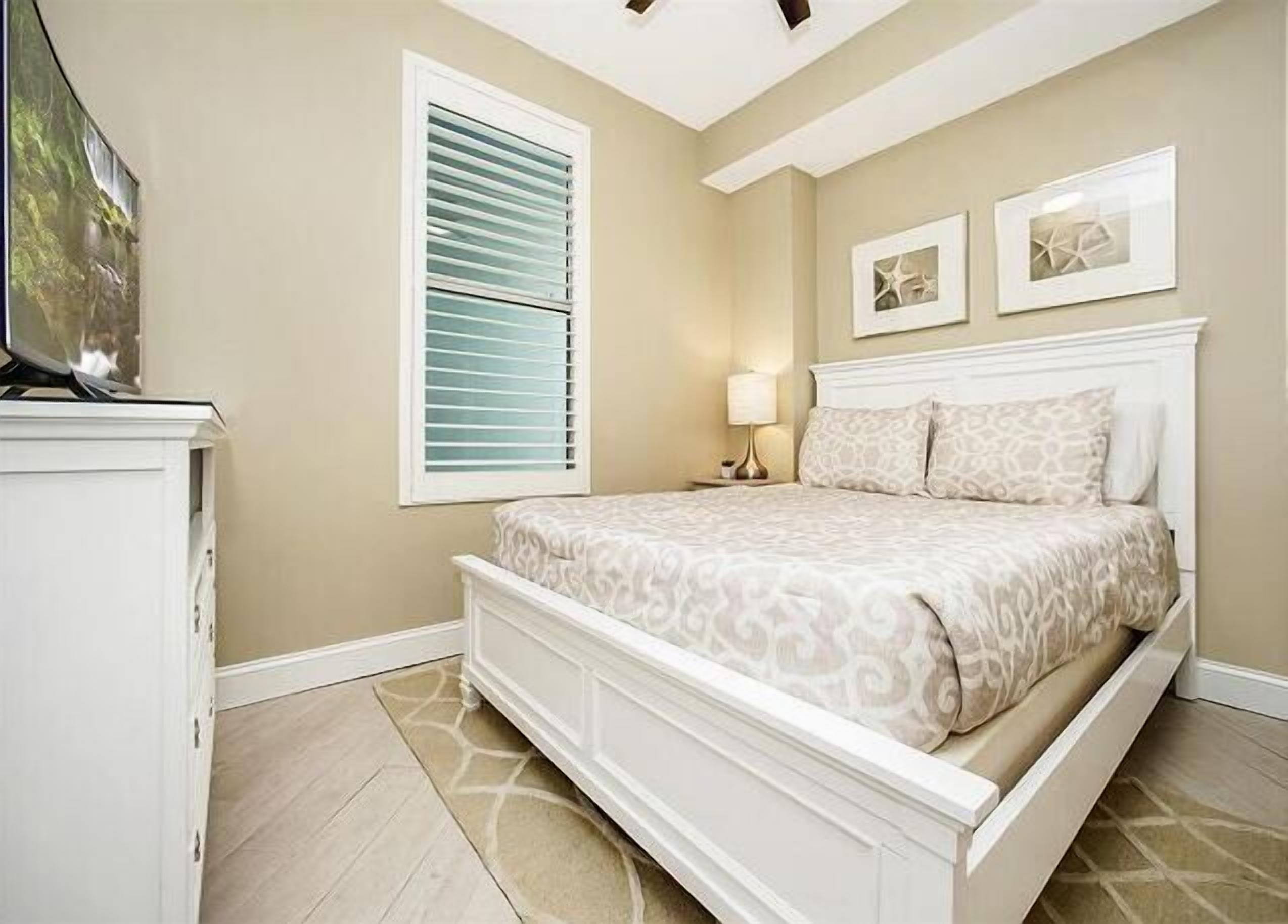 A bedroom with a bed, a nightstand, a lamp, a window with blinds, and artwork on the wall.
