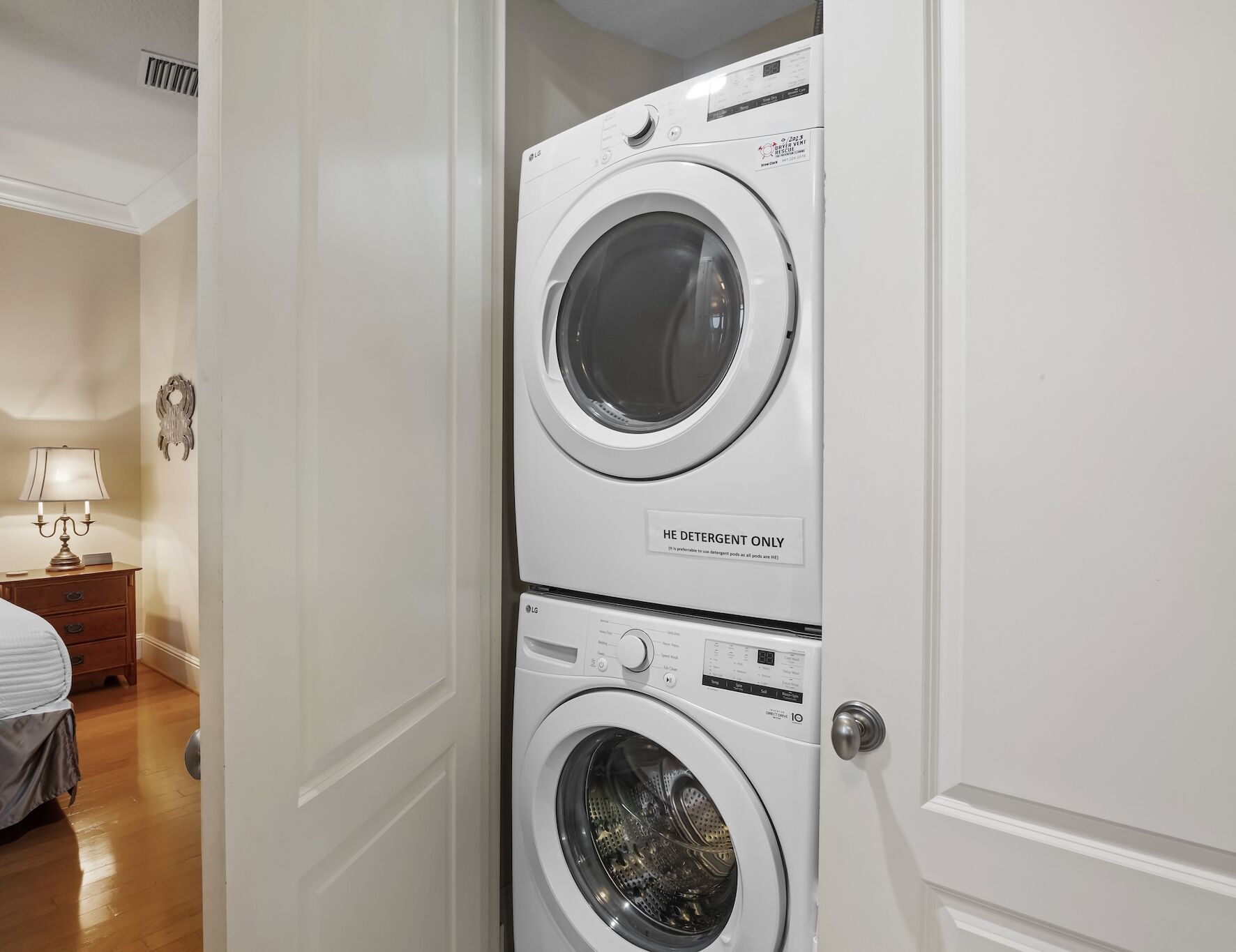 A laundry room with two stacked front-loading washing machines, a wooden nightstand with a lamp, and a bed in the adjacent room.