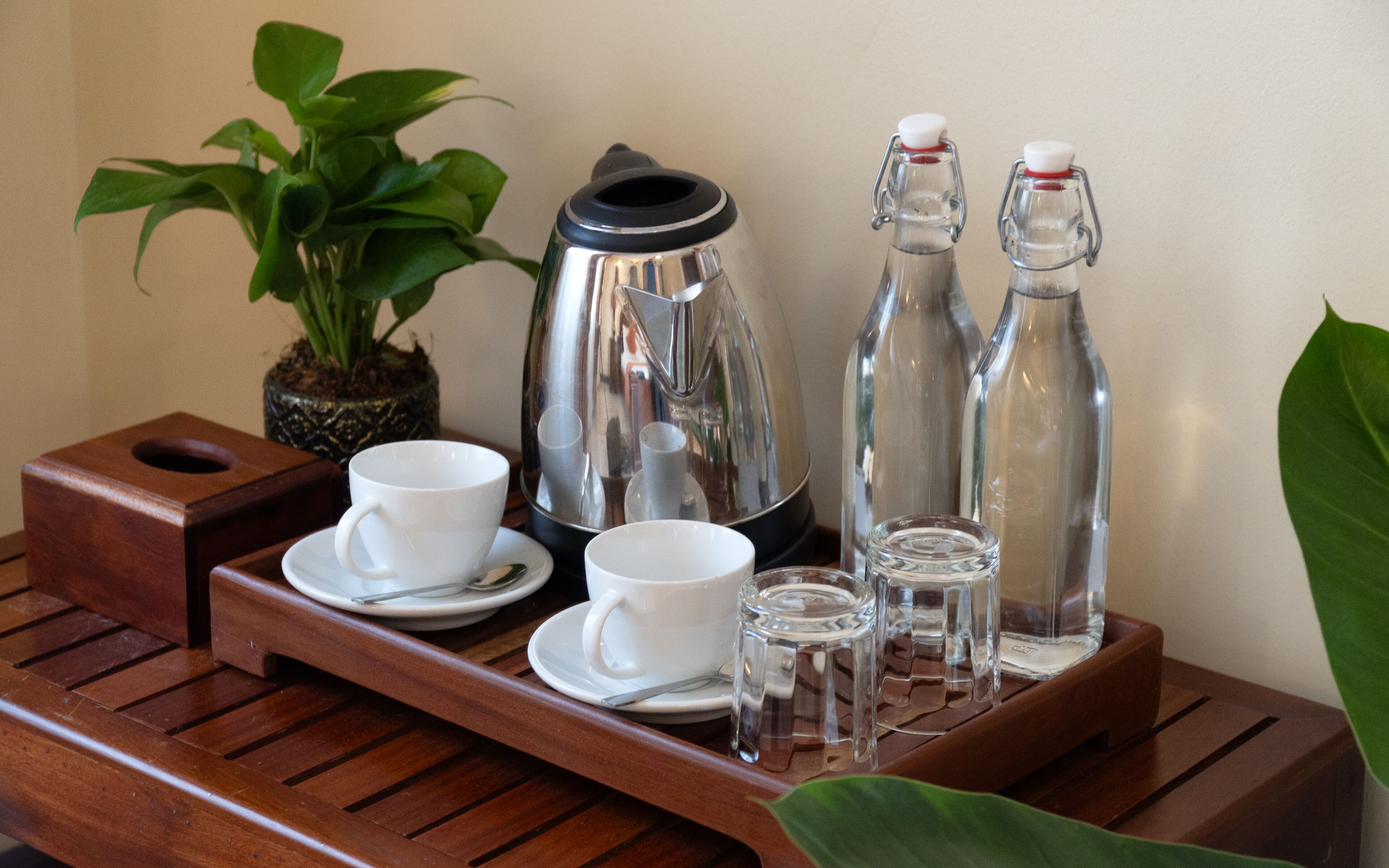 Una bandeja de madera con una cafetera, dos botellas, dos tazas y un vaso sobre una mesa.