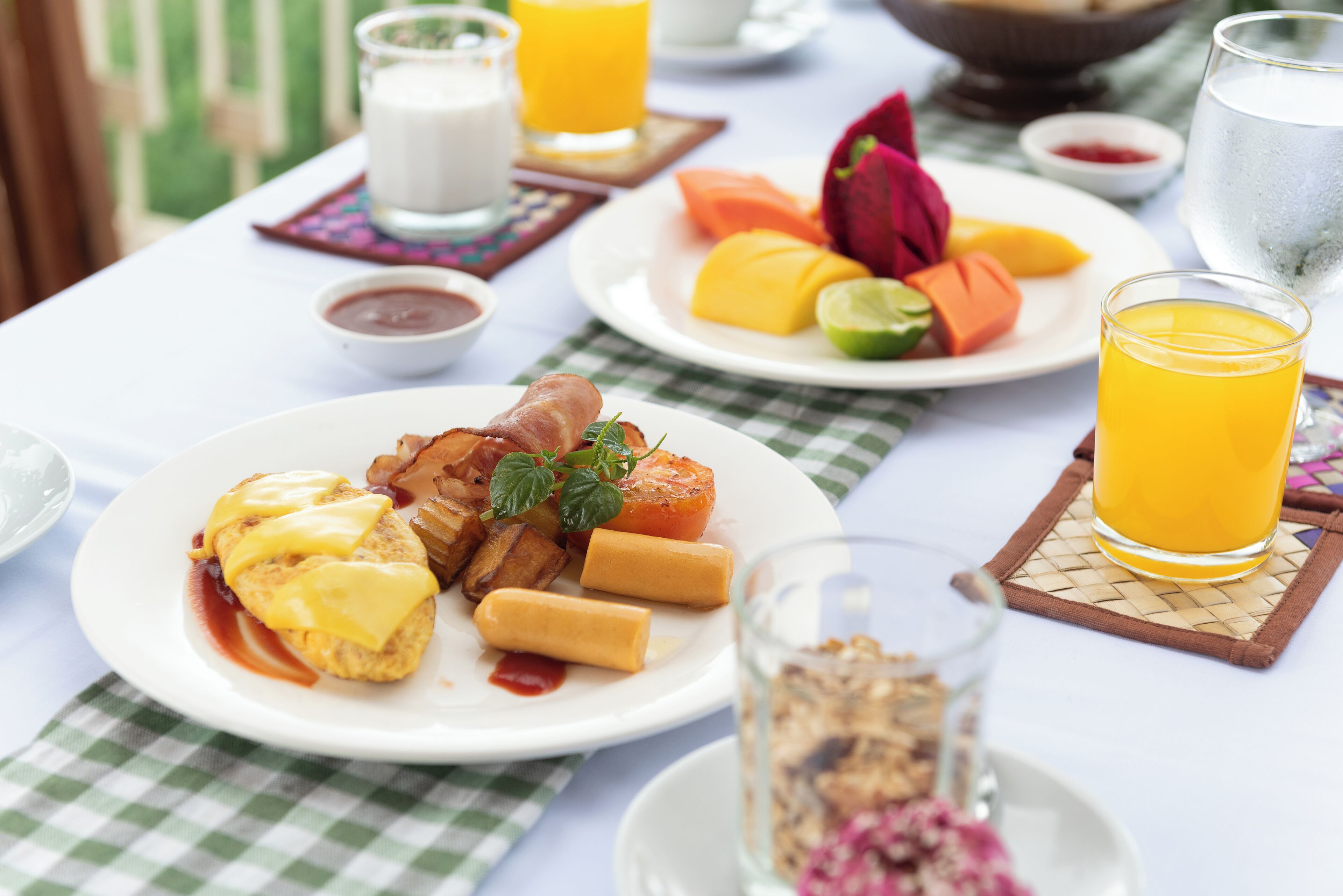 En morgenmadsbordopstilling med tallerkener med æg, bacon og grøntsager, et glas appelsinjuice og en skål med frugt.