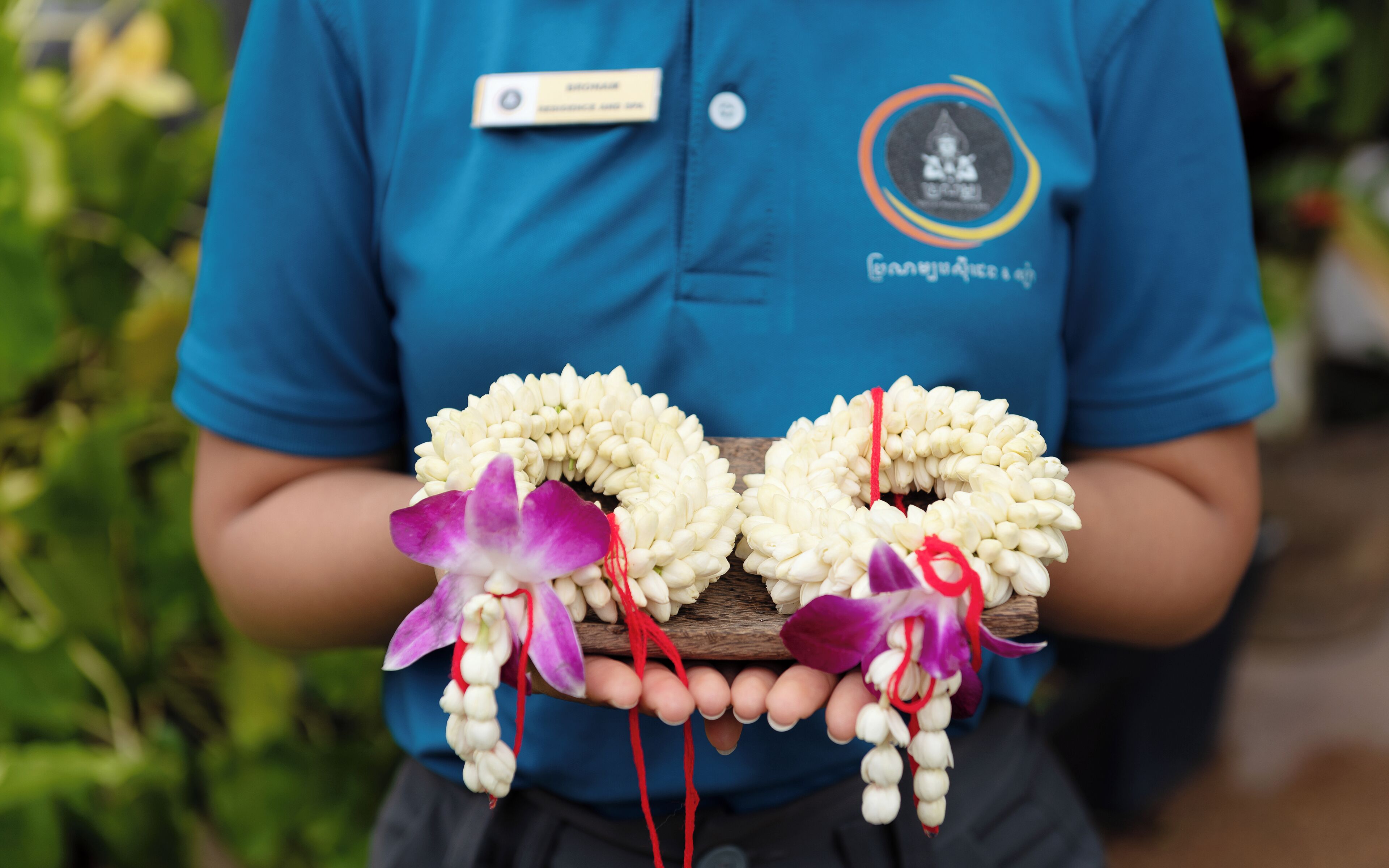 Una persona sostiene dos guirnaldas de flores blancas con orquídeas moradas, vistiendo una camisa azul con una etiqueta de nombre y un logotipo.