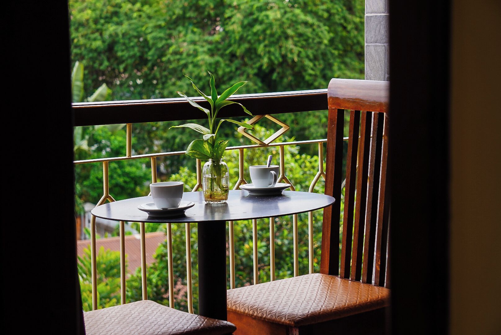 A balcony with a round table, two white cups, a small vase with a plant, and a wooden chair.