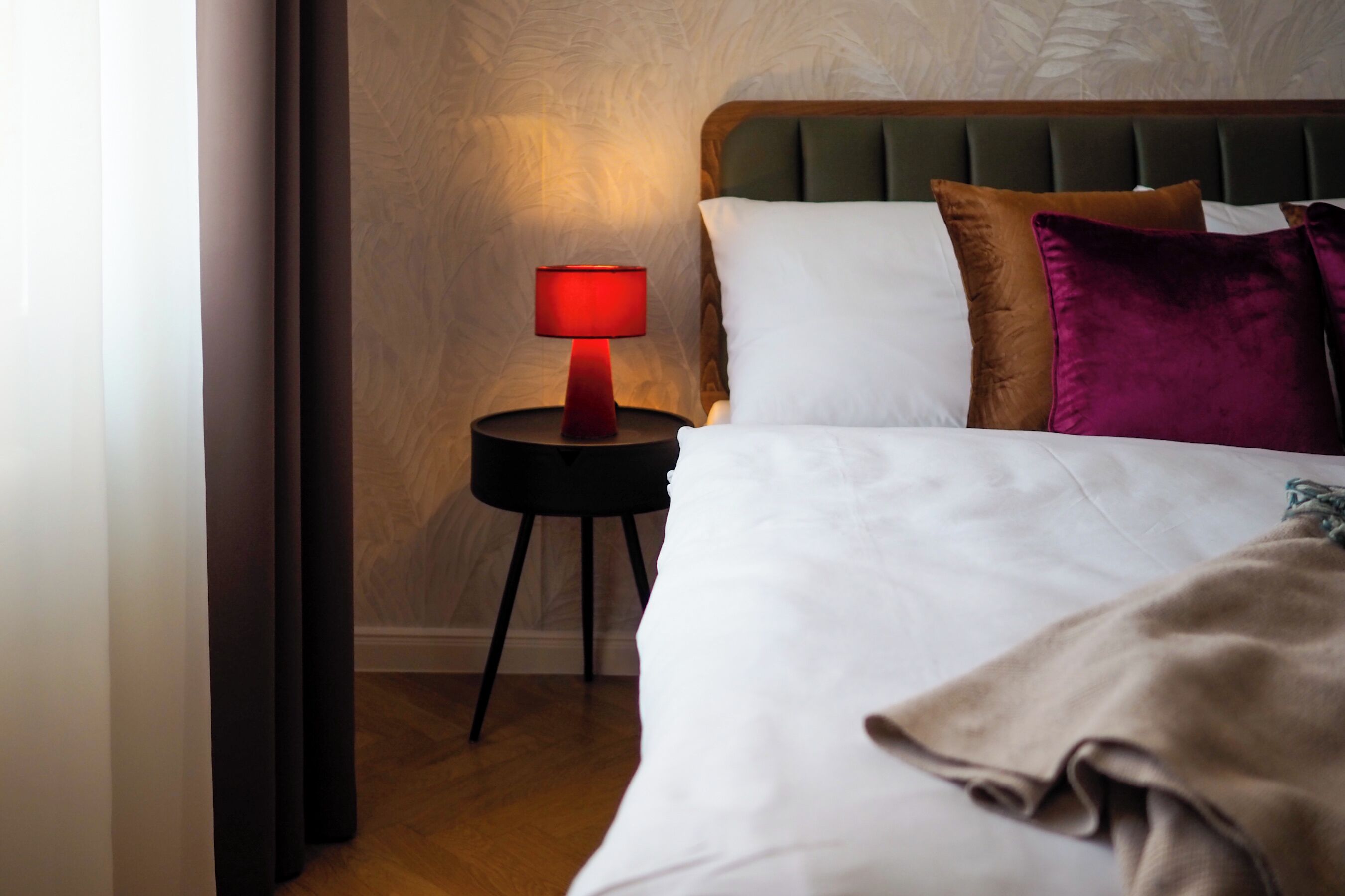 Una cama bien hecha con ropa de cama blanca y marrón, una lámpara roja sobre una mesita de noche negra pequeña y un cabecero tapizado de color verde.