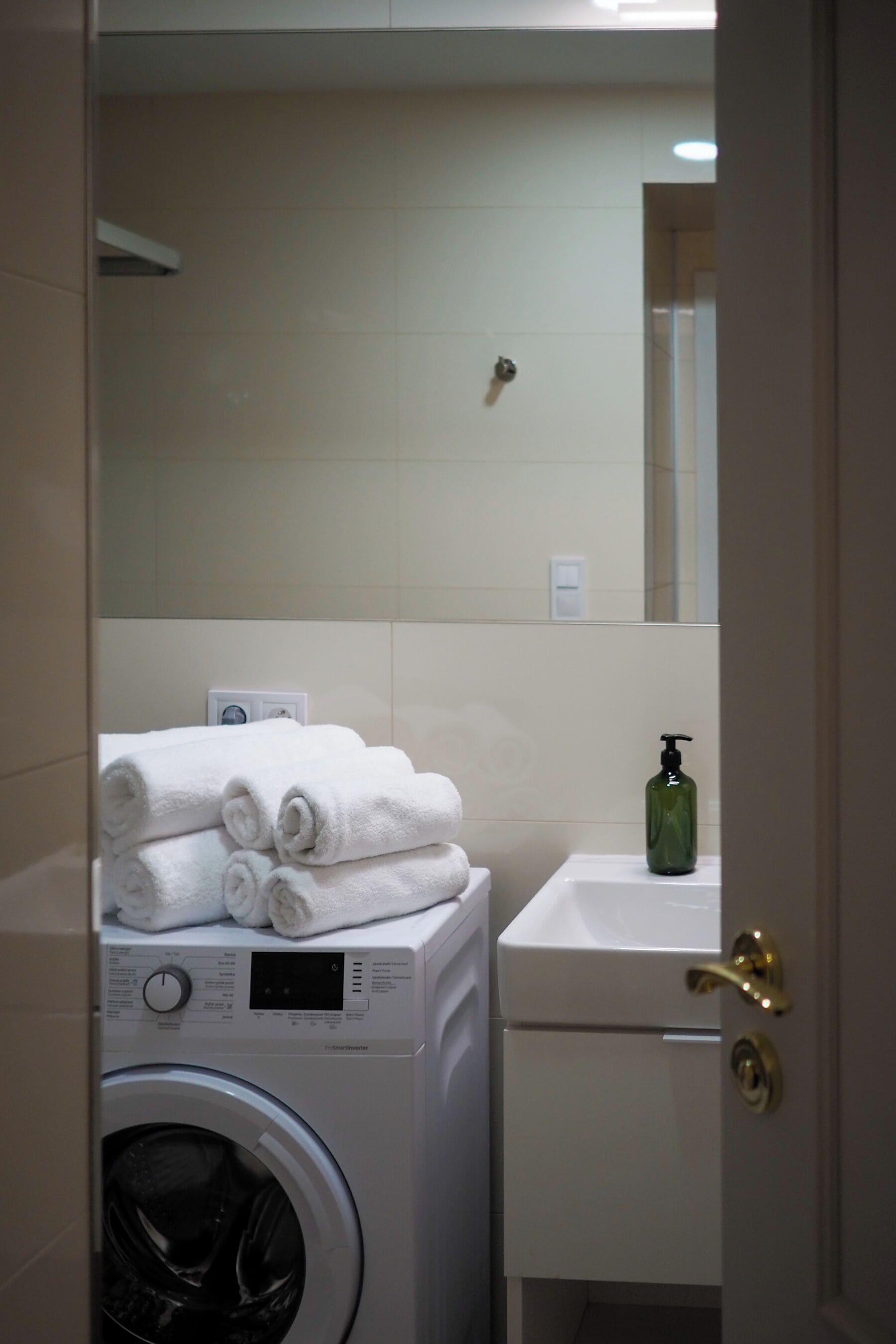 A compact laundry area with a washing machine, a stack of white towels, a green soap dispenser, and a small sink.