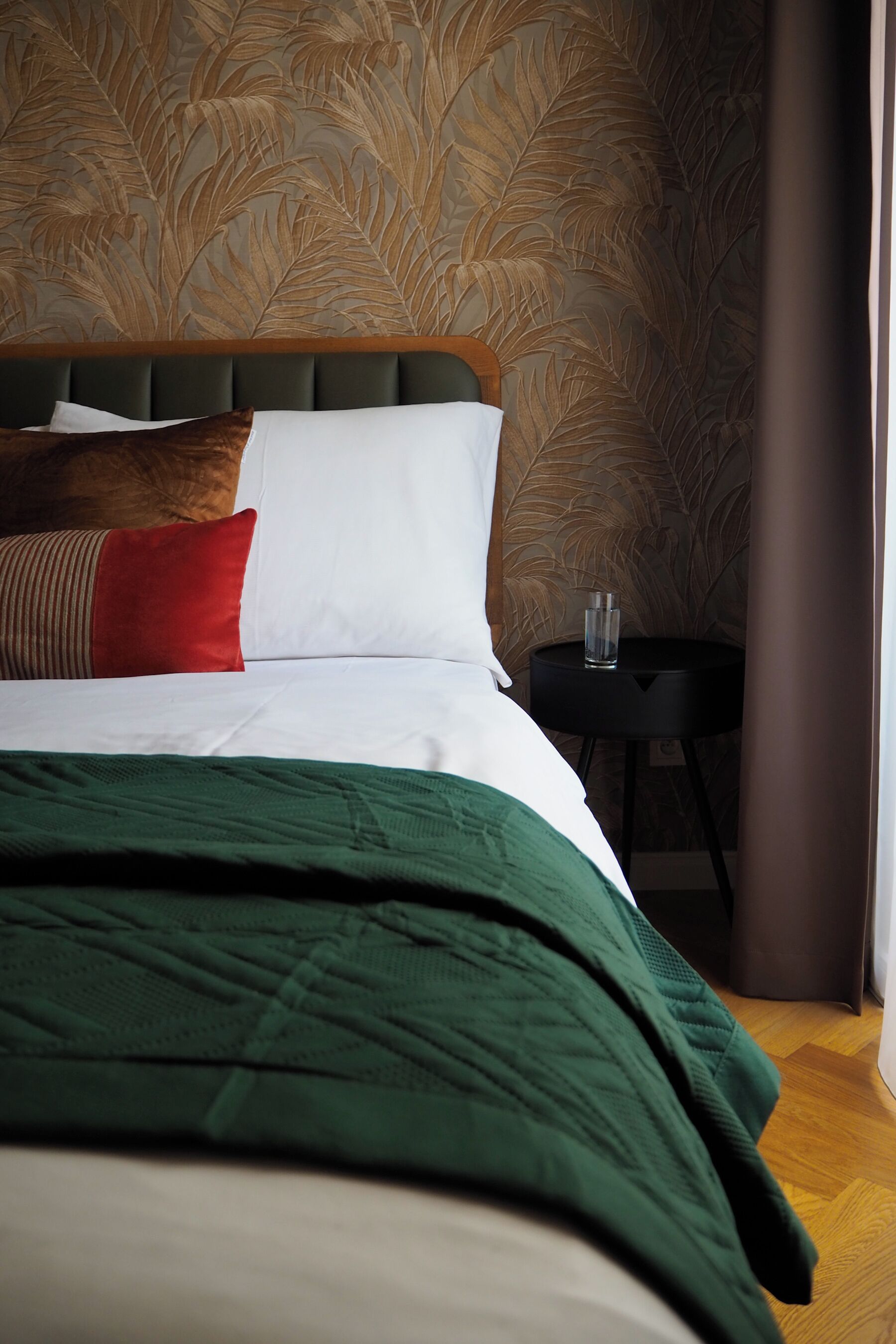 A neatly made bed with a green blanket, white and red pillows, and a wooden headboard. A small black table with a glass of water is beside the bed.