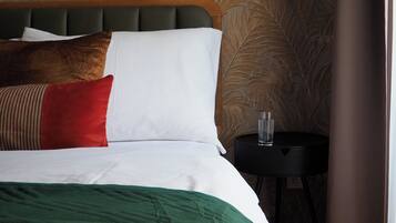 A neatly made bed with a green blanket, white and red pillows, and a wooden headboard. A small black table with a glass of water is beside the bed.