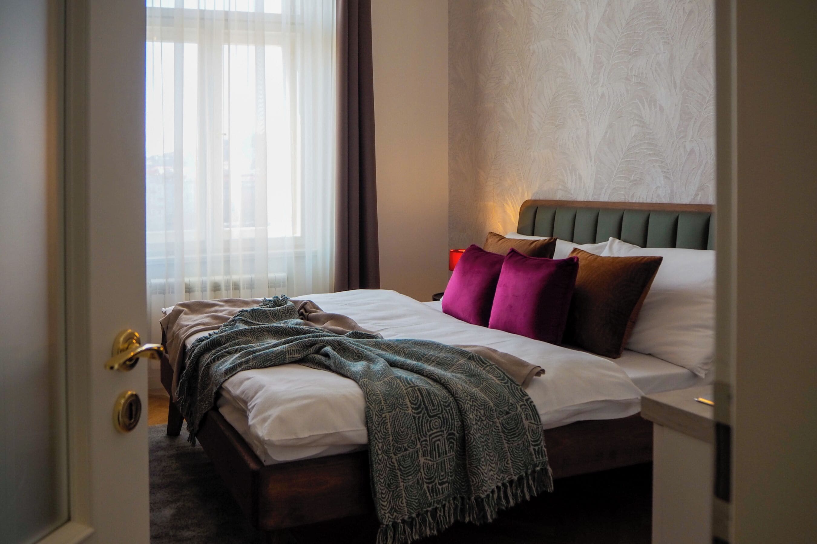 A hotel room with a bed, a nightstand, a window with curtains, and a door with a golden handle.