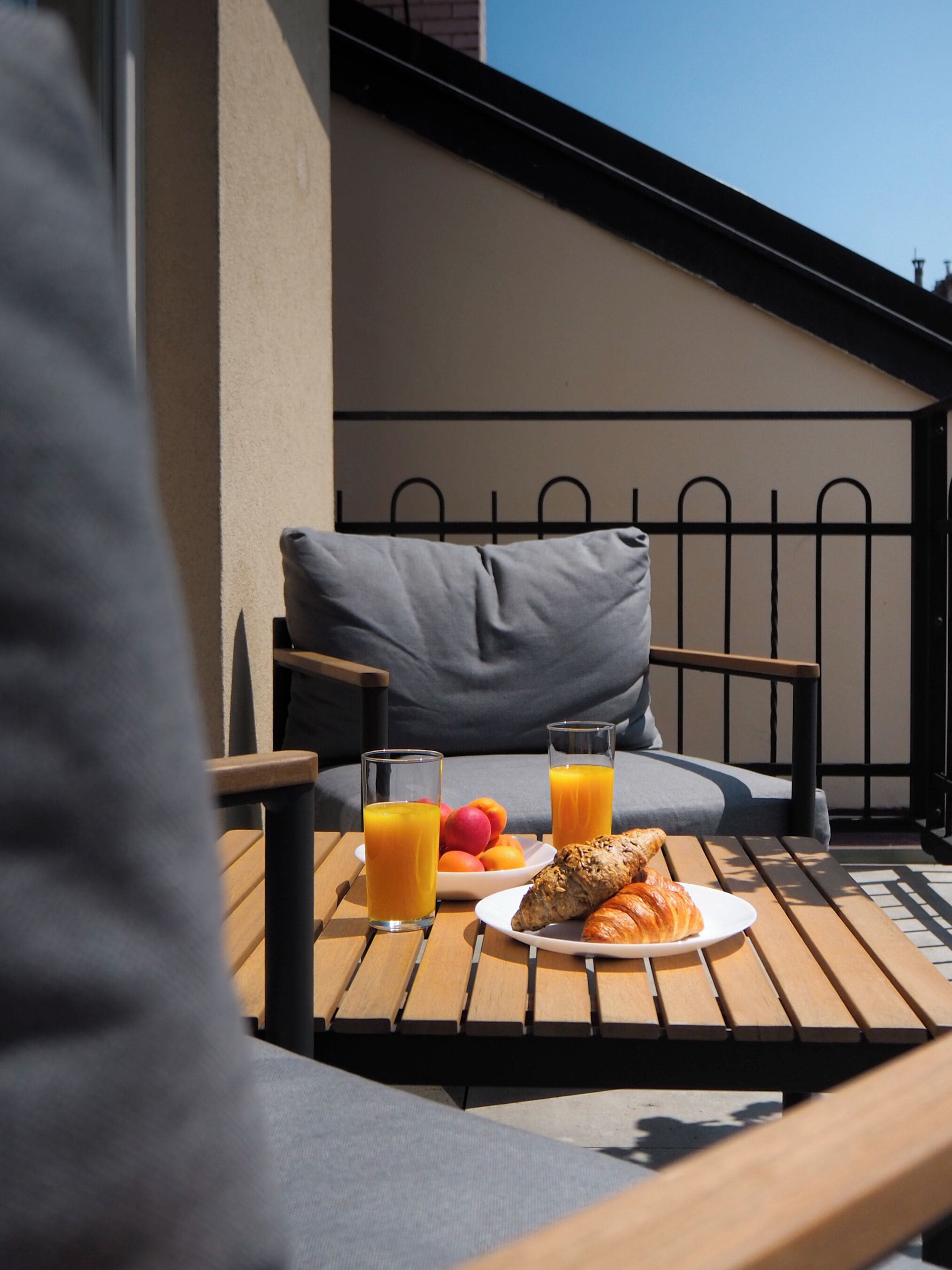 A wooden table with a plate of food, two glasses of orange juice, and a bowl of fruit on an outdoor patio.