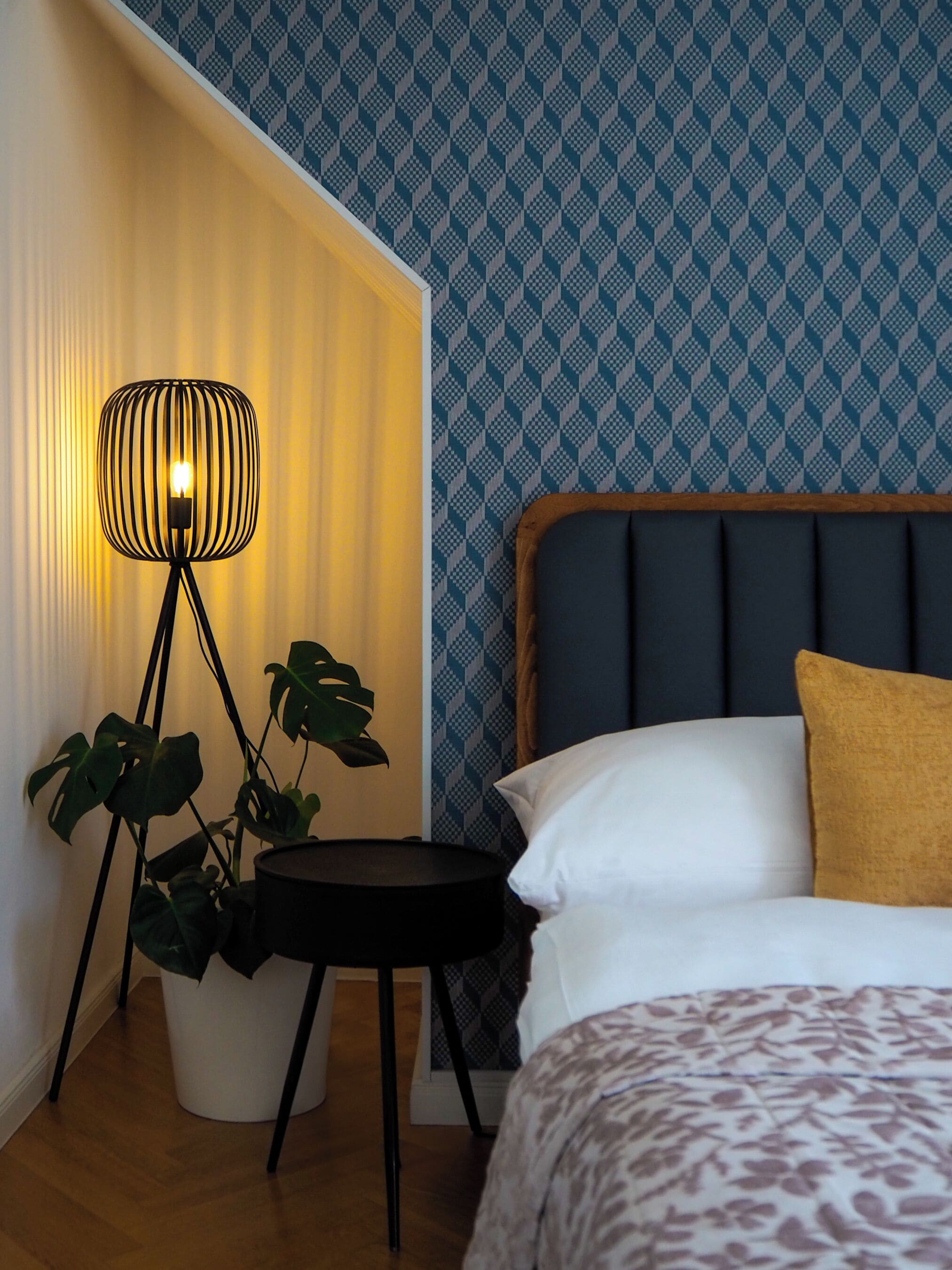 Een slaapkamer met een bed, nachtkastje met lamp, een kamerplant en een behang met patroon.