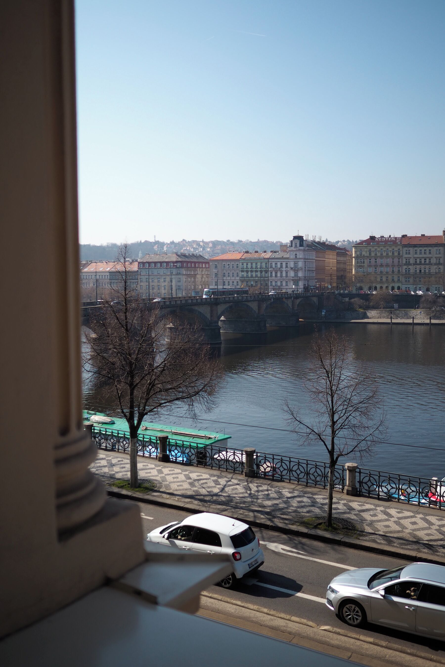 A view of a river with a bridge, buildings, and parked cars.