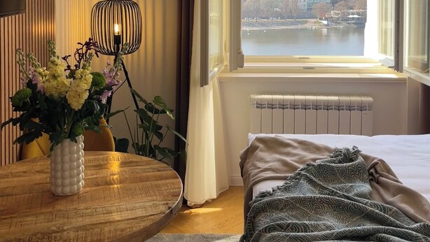 A hotel room with a bed, a table with a vase of flowers, a lamp, and a view of a cityscape through a large window. - Rašínovo nábřeží - Riverside Residence (Prague)